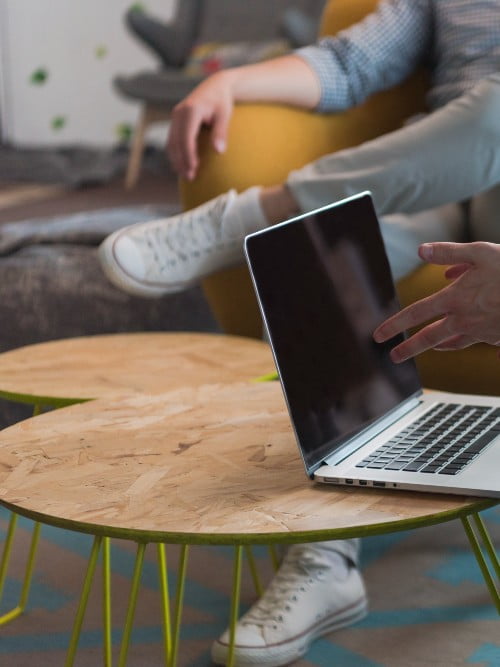 Aufgeklappt es MacBook Pro, dass auf einem Tisch steht. Im Hintergrund sitzt ein Mann auf einem gelben Stuhl.