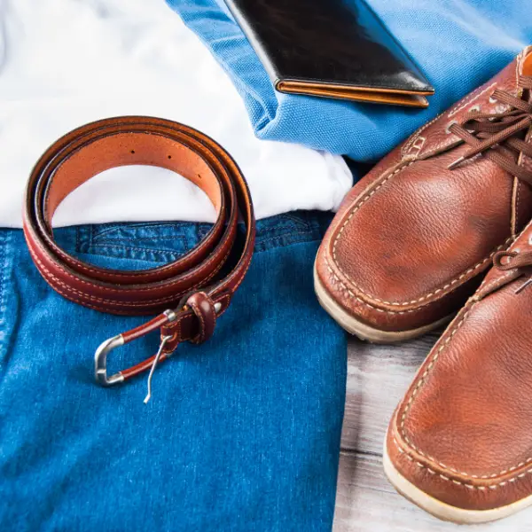 Männer Kleidung die aus einem braunen Gürtel, braunen Schuhen, blaue Jeans, weiße Hemd, blauen T-Shirt und einem schwarzen Portmonee bestehen welches auf einem Holztisch liegt.