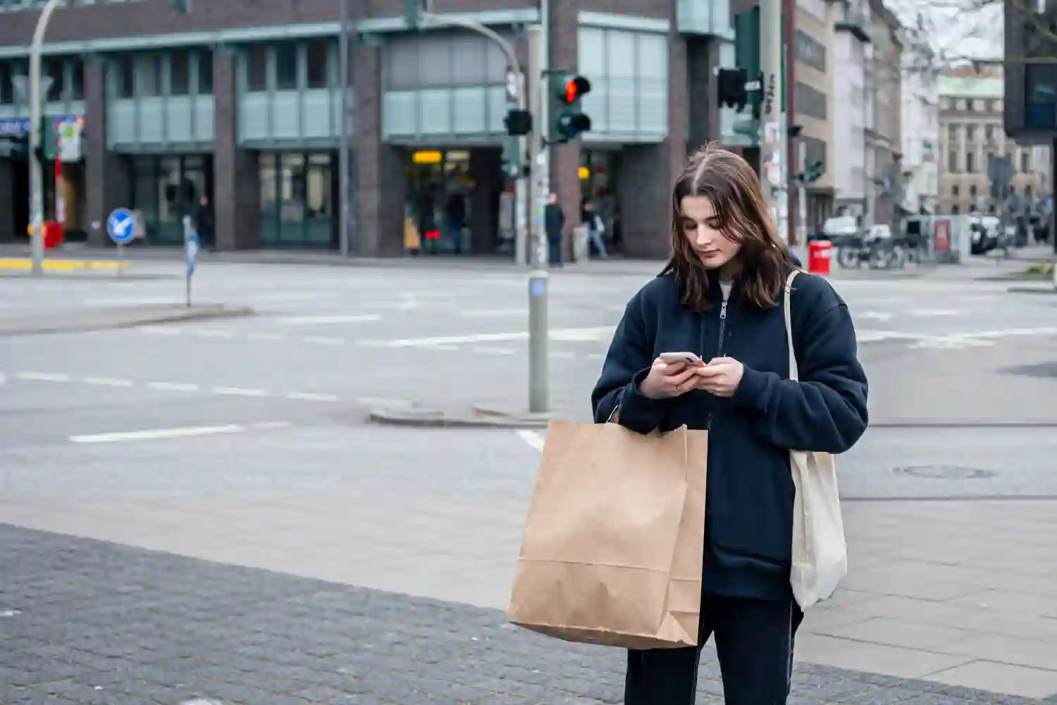 SEO Hannover - Eine Person steht an einer Straßenecke in der Stadt und schaut auf ihr Telefon. Sie hält eine große braune Papiertüte und eine beige Einkaufstasche in der Hand. Im Hintergrund sind eine Ampel und ein Gebäude mit Glasfenstern zu sehen. Local SEO Agentur Hannover.