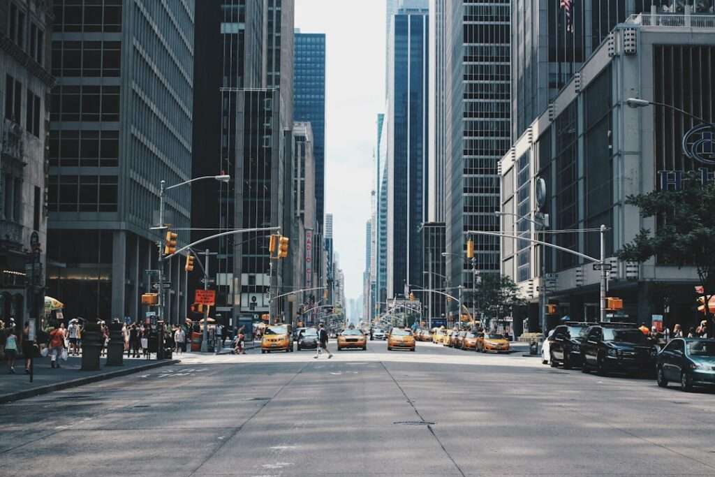 SEO Hannover - Eine belebte Stadtstraße, die auf beiden Seiten von hohen Gebäuden gesäumt ist. Auf der Straße sind gelbe Taxis und Autos unterwegs, und an den Straßenecken haben sich Fußgänger versammelt. Ampeln sind sichtbar und der Himmel ist teilweise bewölkt. Local SEO Agentur Hannover.