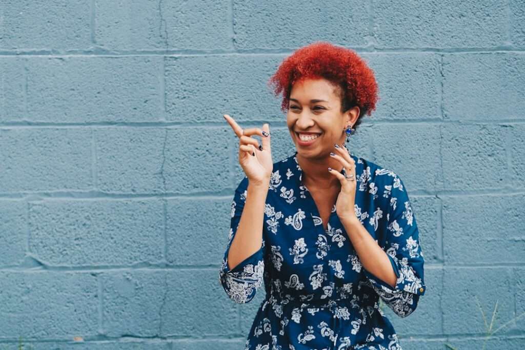 Suchmaschinenoptimierung Hannover - Frau mit lockigem rotem Haar steht lächelnd vor einer blauen Backsteinwand und zeigt mit dem Finger auf etwas. Sie trägt ein blaues Kleid mit weißem Blumenmuster. Ihr fröhlicher Gesichtsausdruck spiegelt die lebendige Essenz von Webdesign Hannover wider. Local SEO Agentur Hannover.