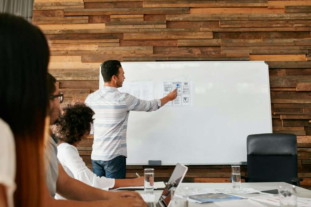 Suchmaschinenoptimierung Hannover - Ein Mann im gestreiften Hemd präsentiert drei sitzenden Kollegen im Büro einer SEO Agentur Hannover Designskizzen auf einem Whiteboard. Eine Person benutzt einen Laptop, eine andere schreibt in ein Notizbuch. Die Wand hinter dem Whiteboard ist mit einem Holzpaneel gestaltet.
 Local SEO Agentur Hannover.