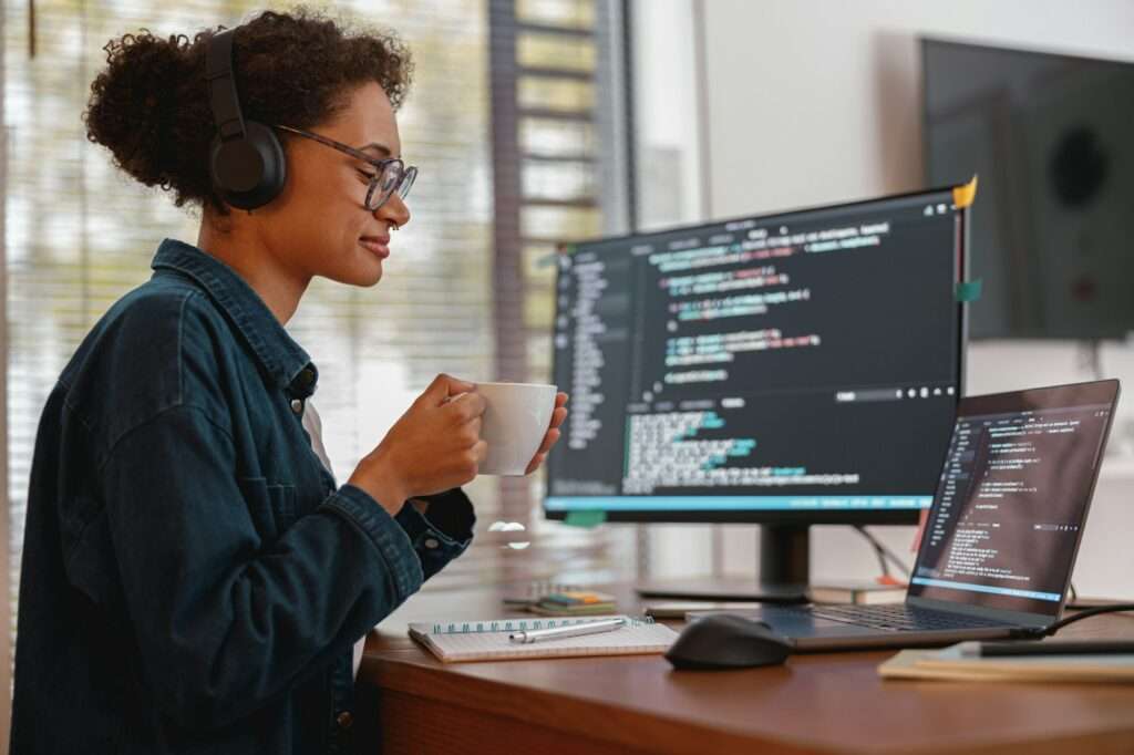 SEO Hannover - Ein Webdesigner in Hannover sitzt mit Kopfhörern an einem Schreibtisch und hält eine Tasse vor den Mund. Vor ihm steht ein Computerbildschirm, auf dem Codezeilen angezeigt werden. Daneben stehen ein weiterer Laptop und ein offenes Notizbuch. Im Hintergrund sind ein Fenster mit Jalousien und ein an der Wand montierter Fernseher zu sehen. Local SEO Agentur Hannover.