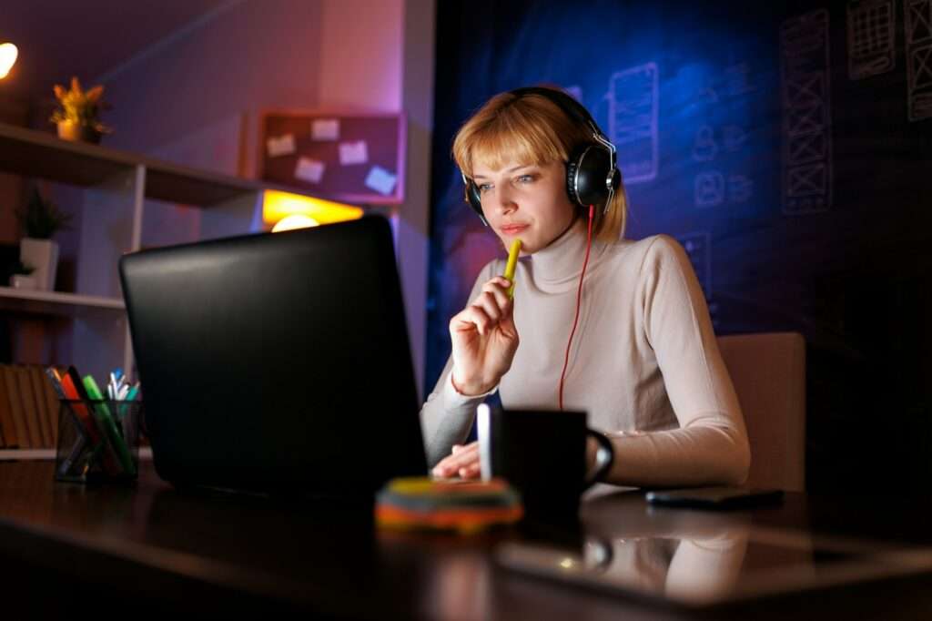 Suchmaschinenoptimierung Hannover - Eine Person mit blonden Haaren, Kopfhörern und einem hellen Rollkragenpullover sitzt an einem Schreibtisch, blickt konzentriert auf einen Laptop-Bildschirm und hält einen Stift vor den Mund. Der Arbeitsplatz des Mitarbeiters der Webagentur Hannover besteht aus einer Tafelwand, einem Regal mit einigen Gegenständen und einer Tasse auf dem Schreibtisch. Local SEO Agentur Hannover.