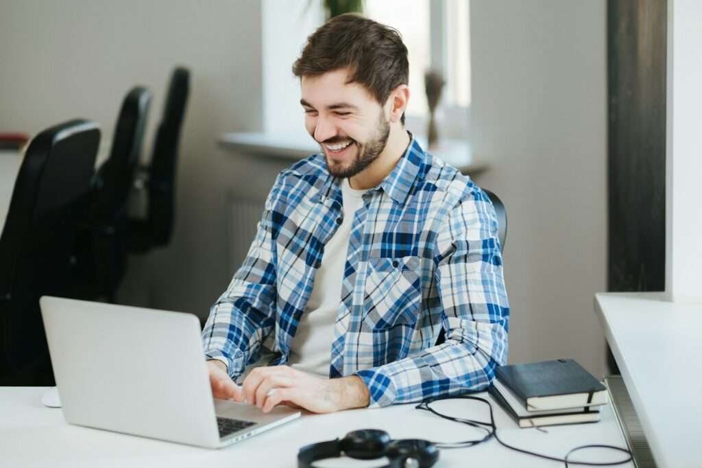 Suchmaschinenoptimierung Hannover - Ein Mann in einem blau-weiß karierten Hemd sitzt lächelnd an einem Schreibtisch und tippt auf einem Laptop. Auf dem Schreibtisch neben ihm liegen zwei geschlossene Notizbücher und ein Paar Kopfhörer. Er scheint sich in einem hellen, modernen Büroraum einer SEO Agentur Hannover zu befinden. Local SEO Agentur Hannover.