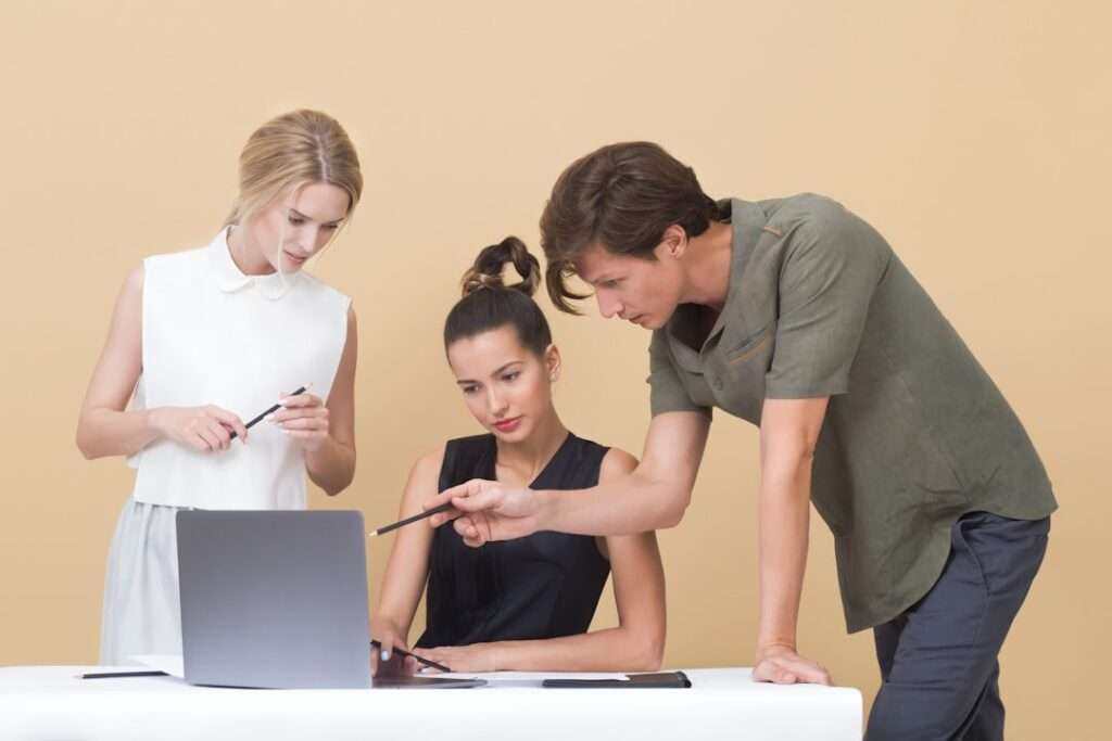 Suchmaschinenoptimierung Hannover - Drei Personen arbeiten gemeinsam an einem Laptop. Eine Frau steht links und hält einen Stift, eine andere Frau sitzt in der Mitte und schaut auf den Bildschirm, und ein Mann steht rechts und zeigt auf den Laptop. Der Hintergrund ist in einem satten Beige gehalten. Local SEO Agentur Hannover.