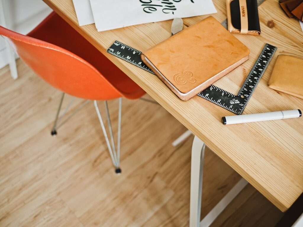 Suchmaschinenoptimierung Hannover - Auf einem Holzschreibtisch liegen ein orangefarbenes Notizbuch, ein schwarzweißes Lineal, ein weißer Marker und einige andere Gegenstände. Neben dem Schreibtisch ist auf einem hellen Holzboden ein orangefarbener Stuhl teilweise sichtbar. Local SEO Agentur Hannover.