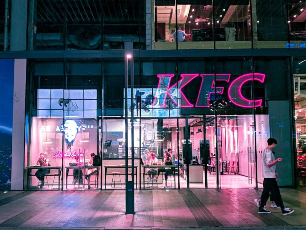 Suchmaschinenoptimierung Hannover - Straßenansicht eines KFC-Restaurants mit einem großen rosa Neonschild mit der Aufschrift „KFC“. Drinnen sieht man Leute, die an Tischen sitzen. Das Gebäude hat eine Glasfront und eine Person geht auf dem Bürgersteig daran vorbei. Es ist Nacht, und die Atmosphäre lässt einen fragen, wie man solche Lokale bei Google bewerten kann. Local SEO Agentur Hannover.