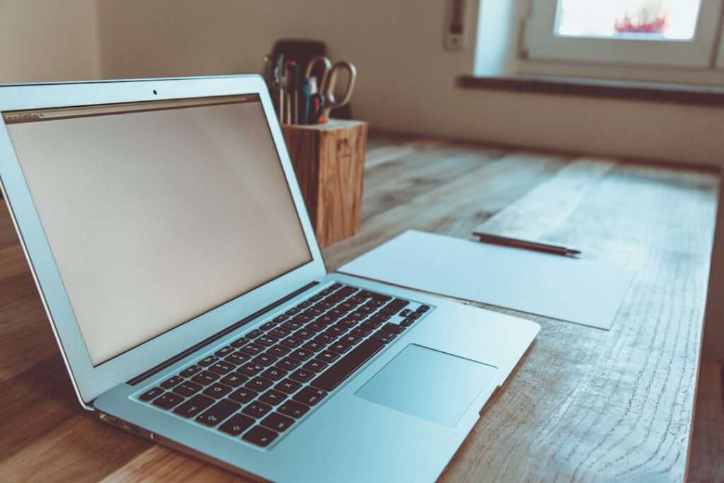 Suchmaschinenoptimierung Hannover - Ein Laptop mit leerem Bildschirm steht neben einem Blatt Papier und einem Stift auf einem Holzschreibtisch und schafft so den perfekten Arbeitsplatz für jeden in der Webagentur Hannover. Im Hintergrund steht ein Behälter mit verschiedenen Stiften und Bleistiften, während natürliches Licht durch das nahe gelegene Fenster strömt. Local SEO Agentur Hannover.