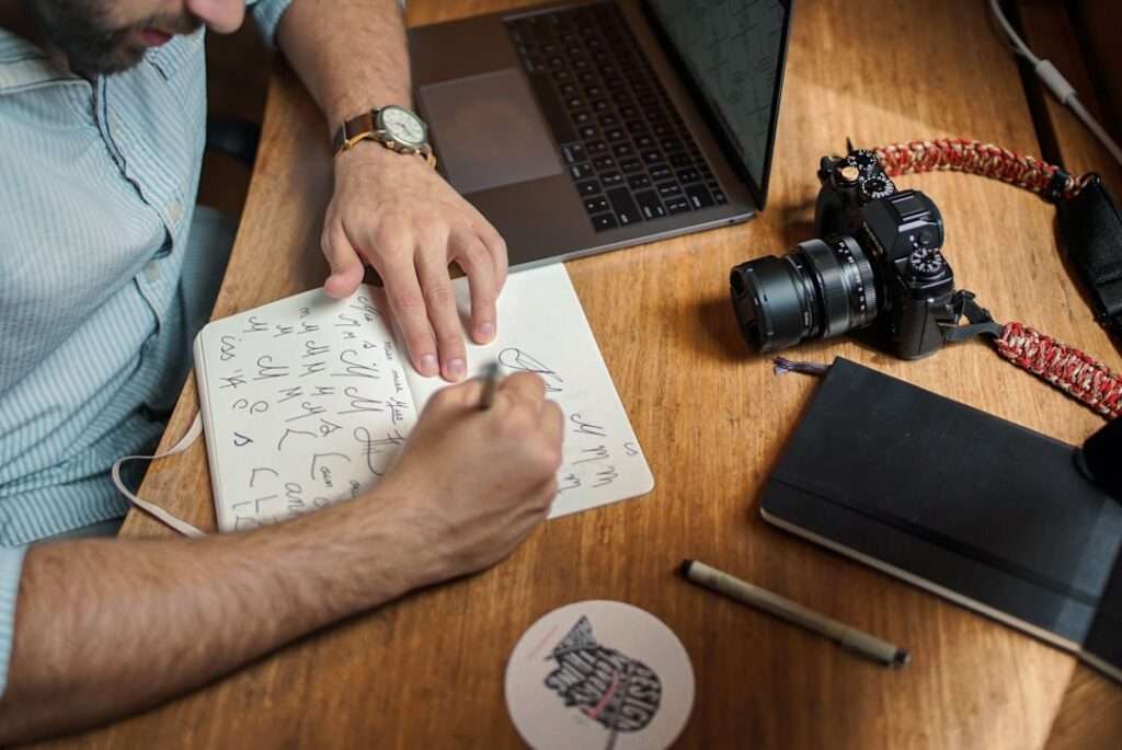 Suchmaschinenoptimierung Hannover - Eine Person schreibt in ein Notizbuch auf einem Holzschreibtisch. Daneben liegen ein Laptop, eine Kamera mit gemustertem Riemen, ein geschlossenes schwarzes Buch, ein Stift und ein runder Untersetzer mit Fischmotiv. Die Person trägt ein gestreiftes Hemd und eine Armbanduhr und skizziert möglicherweise Ideen für das nächste Projekt der Webagentur Hannover. Local SEO Agentur Hannover.