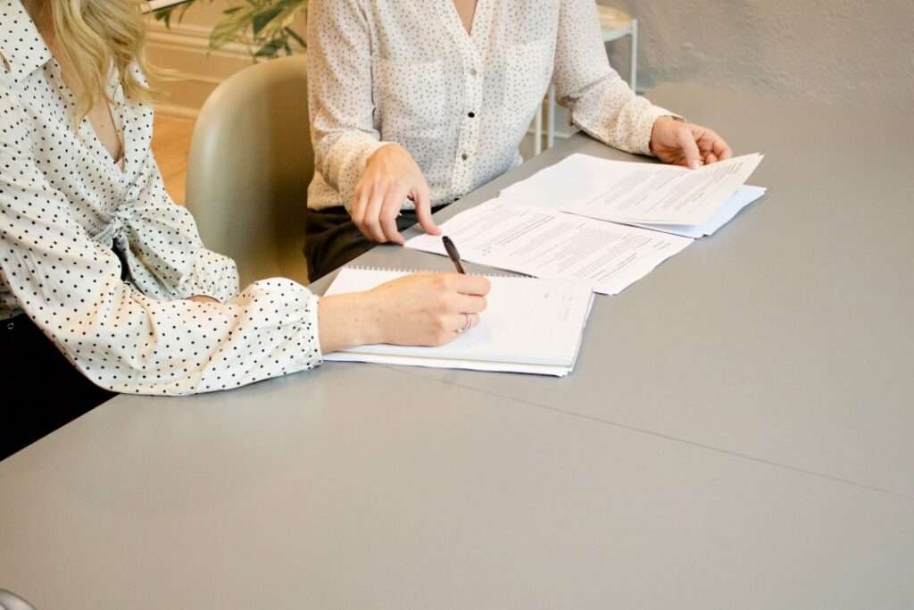 Suchmaschinenoptimierung Hannover - Zwei Personen sitzen an einem mit Dokumenten übersäten Tisch und prüfen die Unterlagen. Beide sind in legerer Business-Kleidung mit weißen, gemusterten Blusen gekleidet, was eine professionelle Büroumgebung suggeriert. Es sieht so aus, als würden sie Strategien für SEO Hannover besprechen. Local SEO Agentur Hannover.