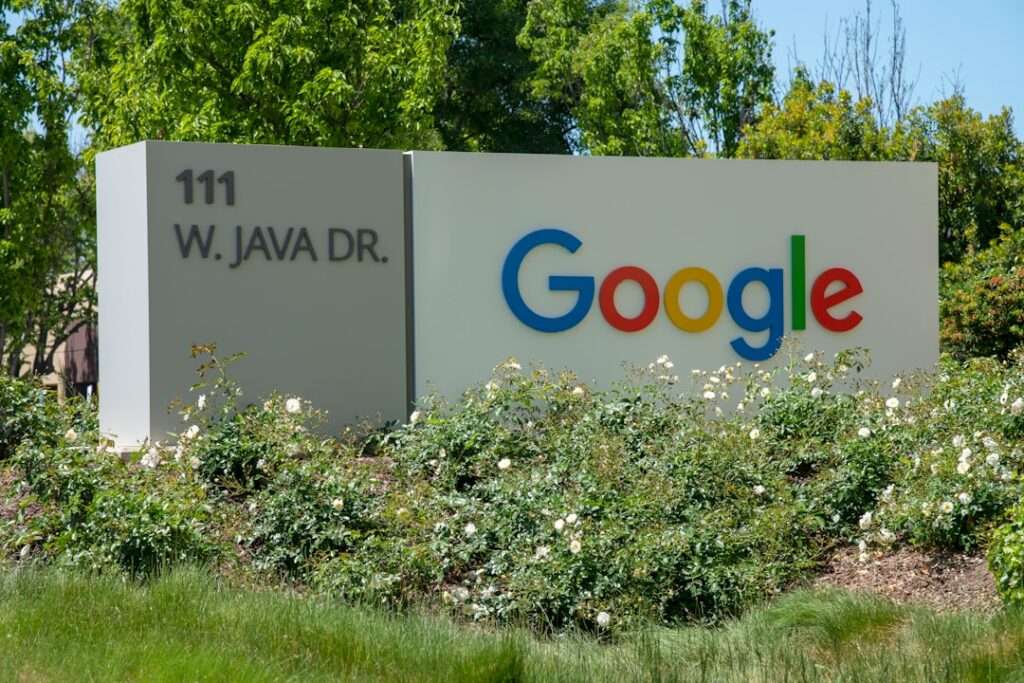 SEO Hannover - Ein großes weißes Schild mit der Adresse „111 W. Java Dr.“ in schwarzer Schrift auf der linken Seite und dem Google-Logo in farbigen Buchstaben auf der rechten Seite, umgeben von Grün und weißen Blumen, ragt hoch auf als Beweis für Innovation – eine perfekte Kulisse für diejenigen, die darüber nachdenken, wie sie mit Google-Bewertungen löschen umgehen sollen. Local SEO Agentur Hannover.