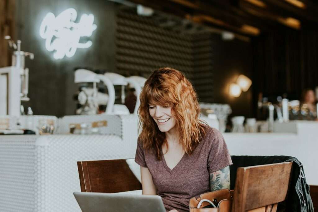 SEO Hannover - Eine Person mit lockigem Haar sitzt an einem Holztisch in einem Café und lächelt auf einen Laptop-Bildschirm. Im Hintergrund ist ein stilvolles Interieur mit einer Neonreklame und einer Kaffeemaschine zu sehen. Local SEO Agentur Hannover.