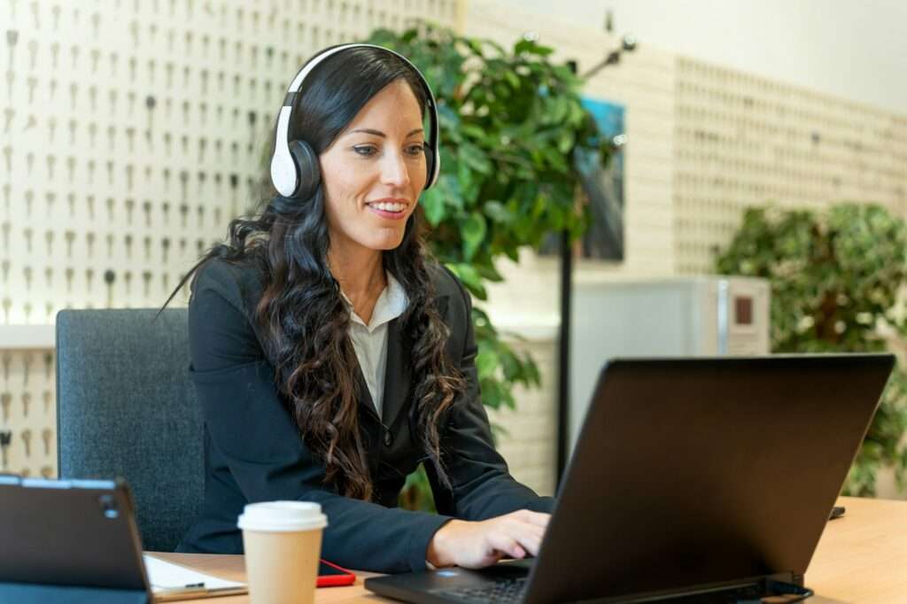 SEO Hannover - Eine Frau mit langen dunklen Haaren, im Business-Outfit, sitzt an einem Schreibtisch mit aufgeklapptem Laptop. Sie trägt weiße Kopfhörer und lächelt, während sie auf den Bildschirm blickt. Auf dem Schreibtisch stehen eine Kaffeetasse, ein Tablet und ein Smartphone, im Hintergrund eine Pflanze – eine typische Szene bei der Webagentur Hannover. Local SEO Agentur Hannover.