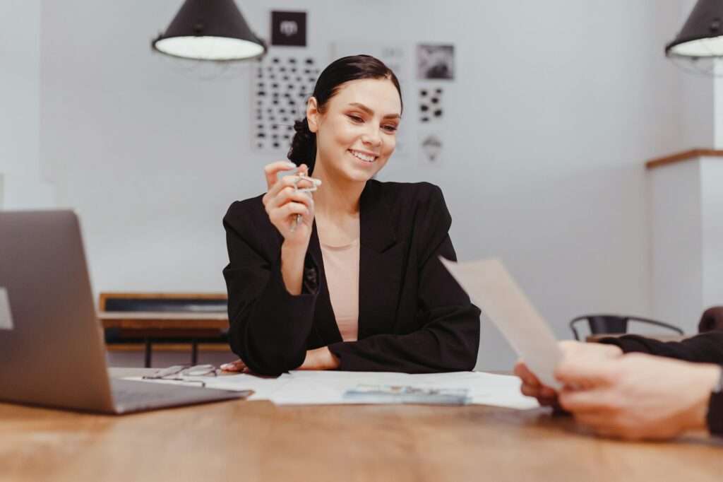 SEO Hannover - Eine Frau mit dunklem Haar und schwarzem Blazer sitzt mit Laptop und Dokumenten an einem Schreibtisch und vertritt die Webagentur Hannover. Sie lächelt und hält einen Stift in der Hand, während sie eine Person am anderen Ende des Tisches ansieht, die ein Blatt Papier in der Hand hält. Im Hintergrund sind eine weiße Wand und eine schwarze Leuchte zu sehen. Local SEO Agentur Hannover.