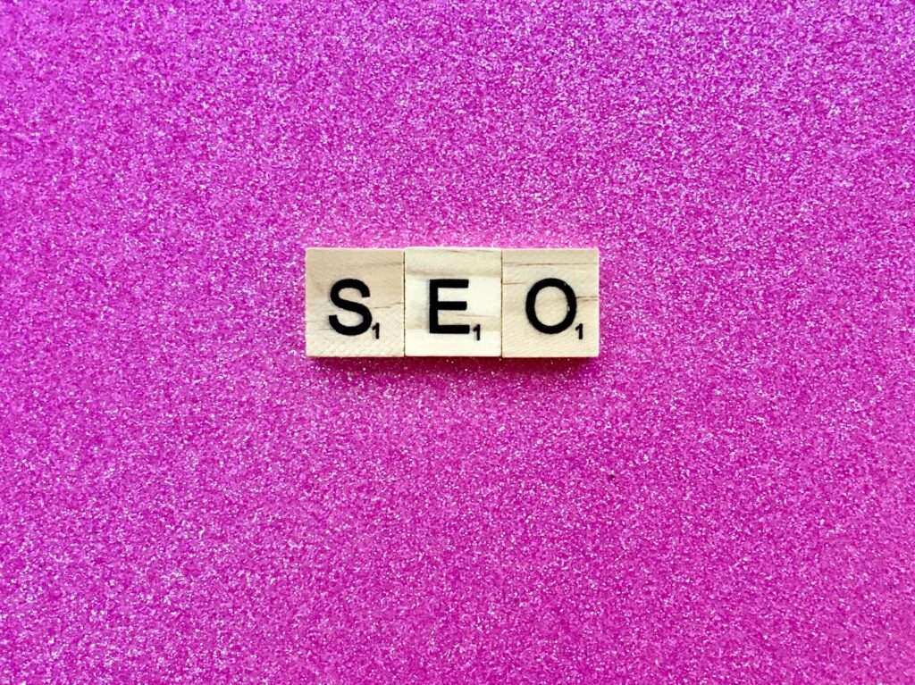 SEO Hannover - Scrabble-Steine mit der Aufschrift „SEO“ liegen nebeneinander auf einem strukturierten, glitzernden rosa Hintergrund. Jeder Stein hat in der unteren rechten Ecke eine kleine Zahl „1“, die seinen Punktwert im Spiel angibt. Local SEO Agentur Hannover.