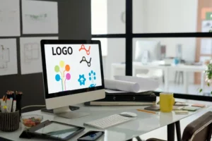 Suchmaschinenoptimierung Hannover - Auf einem Computerbildschirm auf einem Bürotisch sind verschiedene farbenfrohe Logodesigns zu sehen, die ein Webdesignprojekt in Hannover hervorheben. Auf dem Schreibtisch befinden sich außerdem eine Tastatur, eine Maus, eine Kaffeetasse und Büromaterial. Im Hintergrund ist ein weiteres Büro mit einer verschwommenen Sicht auf Möbel und andere Arbeitsplätze zu sehen. Local SEO Agentur Hannover.