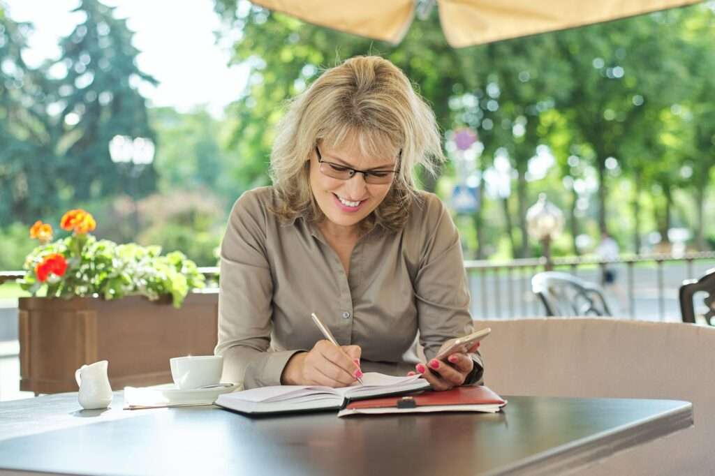 SEO Hannover - Eine Frau mit Brille sitzt an einem Cafétisch im Freien und balanciert gekonnt die Google SEO-Recherche auf ihrem Smartphone, während sie sich Notizen in ihr Notizbuch macht. Eine Kaffeetasse und ein kleiner Krug stehen neben ihr, umgeben von leuchtenden Blumen und Grünpflanzen. Local SEO Agentur Hannover.