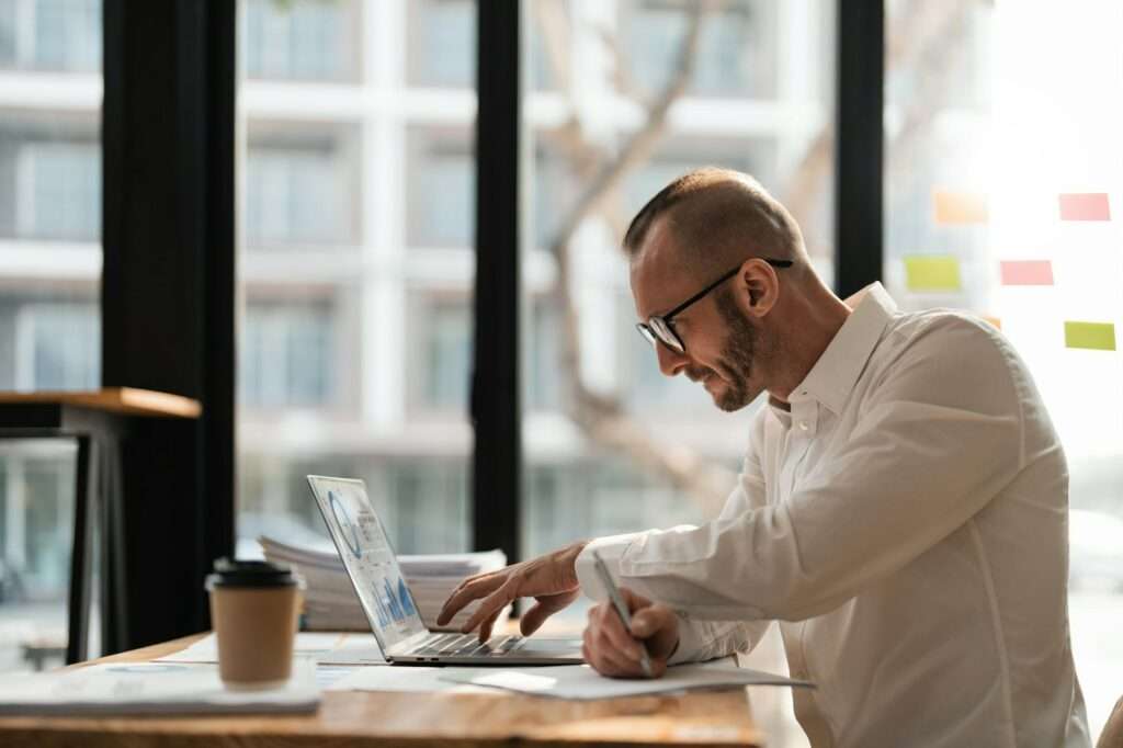 SEO Hannover - Ein Mann arbeitet fleißig an seinem Schreibtisch und nutzt einen Laptop, um Google My Business-Einträge zu optimieren. Er kritzelt Notizen, während er eine Brille und ein frisches weißes Hemd trägt. Eine dampfende Kaffeetasse steht daneben und große Fenster bieten einen Blick auf das mit Haftnotizen beklebte Gebäude draußen. Local SEO Agentur Hannover.