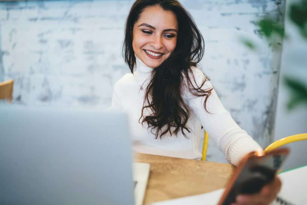 SEO Hannover - Eine Frau mit langen dunklen Haaren und einem weißen Rollkragenpullover sitzt mit einem Laptop an einem Tisch. Sie lächelt in ihr Telefon, vielleicht erfreut über eine Nachricht über die Kosten für Website-Erstellung. Der Hintergrund ist verschwommen, rechts sind eine helle Wand und etwas Grün zu sehen. Local SEO Agentur Hannover.