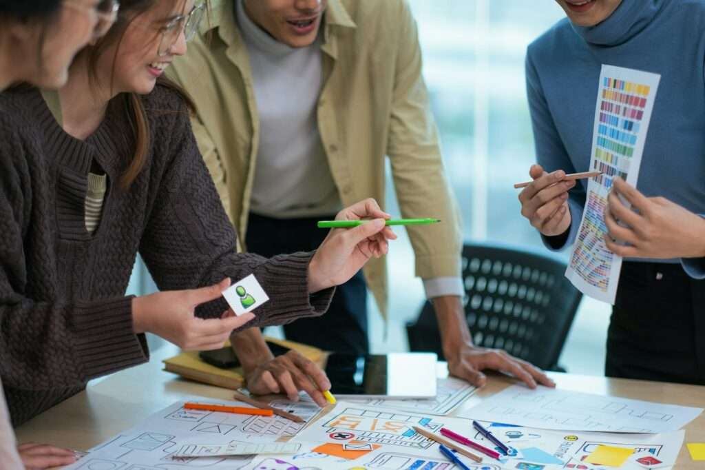 SEO Hannover - Vier Personen arbeiten gemeinsam an einem Tisch, der mit Papieren, Skizzen und Farbmustern bedeckt ist. Einer hält einen grünen Bleistift, während ein anderer eine Farbpalette zeigt. Sie scheinen engagiert und begeistert von dem Projekt zu sein und hoffen, negative Google-Bewertungen zu vermeiden, die die Aufnahme ihrer Arbeit beeinträchtigen könnten. Local SEO Agentur Hannover.