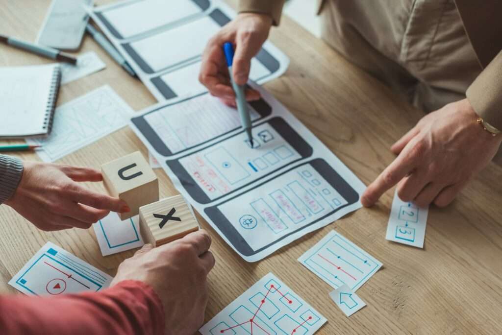 SEO Hannover - Menschen arbeiten gemeinsam an einem Tisch und skizzieren die Benutzeroberfläche einer mobilen App. Zwei Hände halten Holzklötze mit der Aufschrift „U“ und „X“, während eine andere Hand mit einem Stift auf die Skizzen zeigt. Auf dem Tisch liegen verschiedene Designelemente verstreut. Local SEO Agentur Hannover.