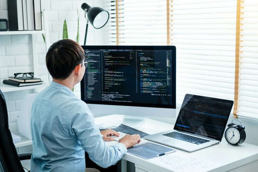 SEO Hannover - Eine Person sitzt an einem Schreibtisch und ist in das Coden auf einem Computermonitor vertieft. In der Nähe zeigt ein Laptop ebenfalls Code an, während ein Wecker und eine Lampe den Arbeitsplatz vervollständigen. Der Raum verfügt über weiße Ziegelwände und große Fenster mit Jalousien – eine perfekte Umgebung, um über die Kosten für die Website-Erstellung nachzudenken. Local SEO Agentur Hannover.