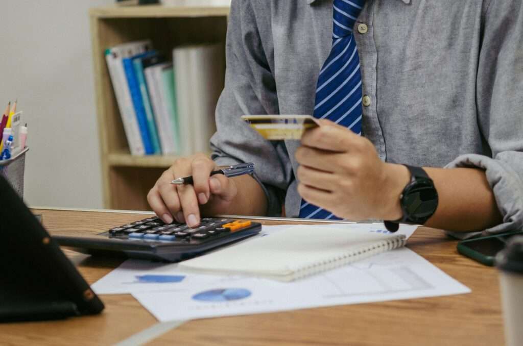 SEO Hannover - Eine Person in grauem Hemd und blau gestreifter Krawatte sitzt an einem Schreibtisch, hält eine Kreditkarte in der Hand und berechnet die Kosten für die Website-Erstellung. Finanzdokumente, ein Notizbuch und ein Tablet liegen auf dem gesamten Arbeitsplatz verteilt. Local SEO Agentur Hannover.