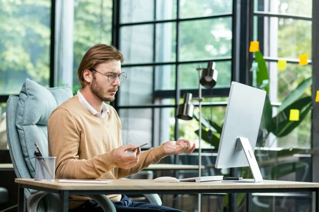 SEO Hannover - Ein Mann mit Brille sitzt auf einem Bürostuhl an einem Schreibtisch, hält einen Stift in der Hand und gestikuliert, während er aufmerksam die Google SEO-Daten auf seinem Computerbildschirm untersucht. Große Fenster mit flatternden Haftnotizen fangen das natürliche Licht ein und eine Pflanze verleiht der Szene einen Hauch von Grün. Local SEO Agentur Hannover.