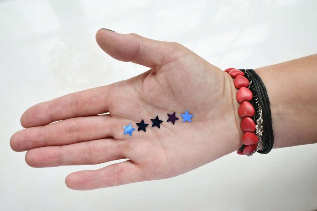 SEO Hannover - Eine mit mehreren Armbändern geschmückte Hand hält fünf kleine, bunte Konfettistücke in Sternform in der Handfläche. Die Sterne sind blau und lila und heben sich von einer schlichten, hellen Oberfläche ab, was an ein festliches und hoch bewertetes Google-Bewertungsbild erinnert. Local SEO Agentur Hannover.