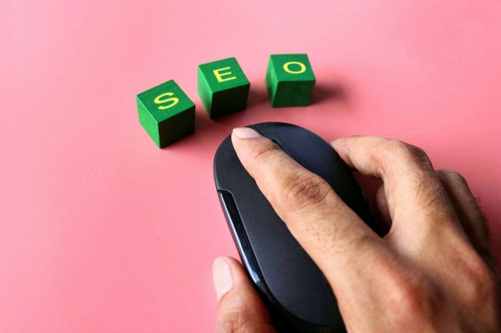 SEO Hannover - Eine Hand bedient eine schwarze Computermaus auf rosa Hintergrund, während darüber grüne Blöcke in gelben Buchstaben das Wort „SEO“ buchstabieren und so die Expertise der besten SEO-Werbeagentur in Hannover symbolisieren. Local SEO Agentur Hannover.