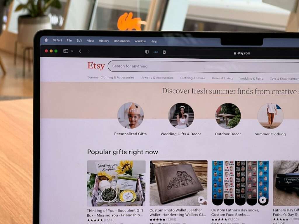 SEO Hannover - Auf einem Laptop-Bildschirm wird die Etsy-Homepage angezeigt, auf der „Momentan beliebte Geschenke“ zu sehen sind, darunter Schätze wie eine saftige Geschenkbox und personalisierte Geldbörsen. Der Safari-Browser, ein unverzichtbares Tool für jede Webdesign-Agentur, zeigt offene Tabs vor einer leicht verschwommenen Pflanze im Hintergrund. Local SEO Agentur Hannover.