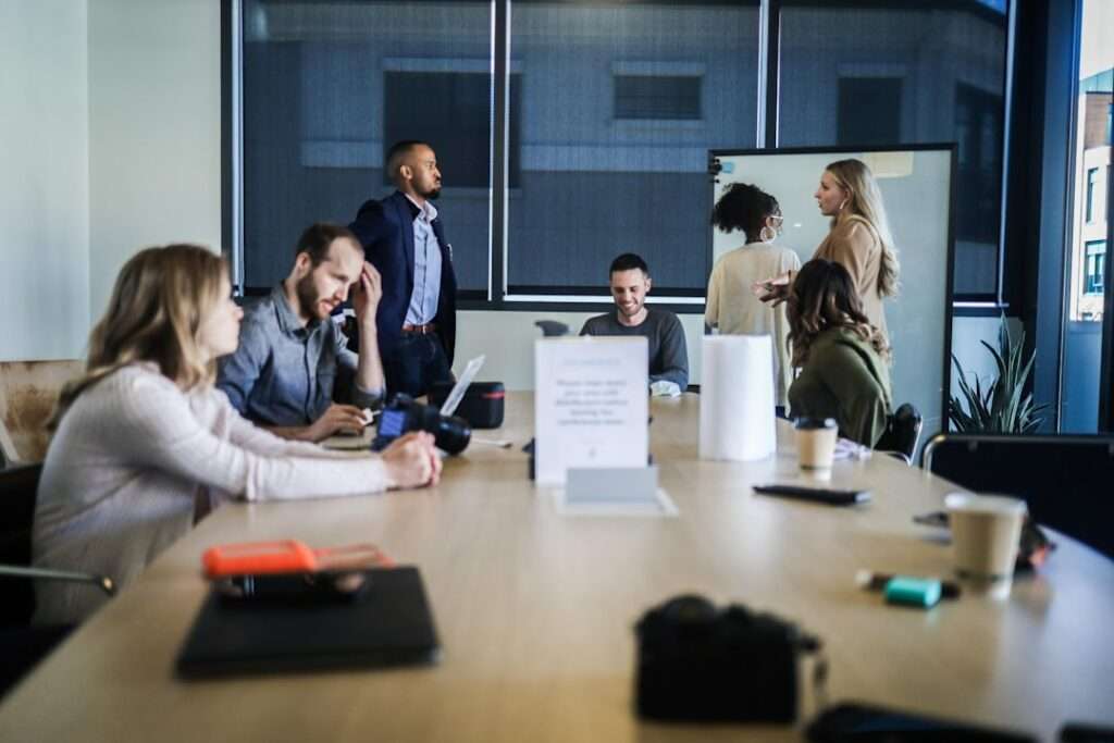 SEO Hannover - Eine Gruppe von Menschen hat sich in einem Konferenzraum versammelt. Einige sitzen mit Laptops und Unterlagen an einem langen Tisch und diskutieren Google-SEO-Strategien, während andere neben einem Whiteboard stehen und sich unterhalten. Der Raum hat große Fenster mit teilweise geschlossenen Jalousien. Local SEO Agentur Hannover.