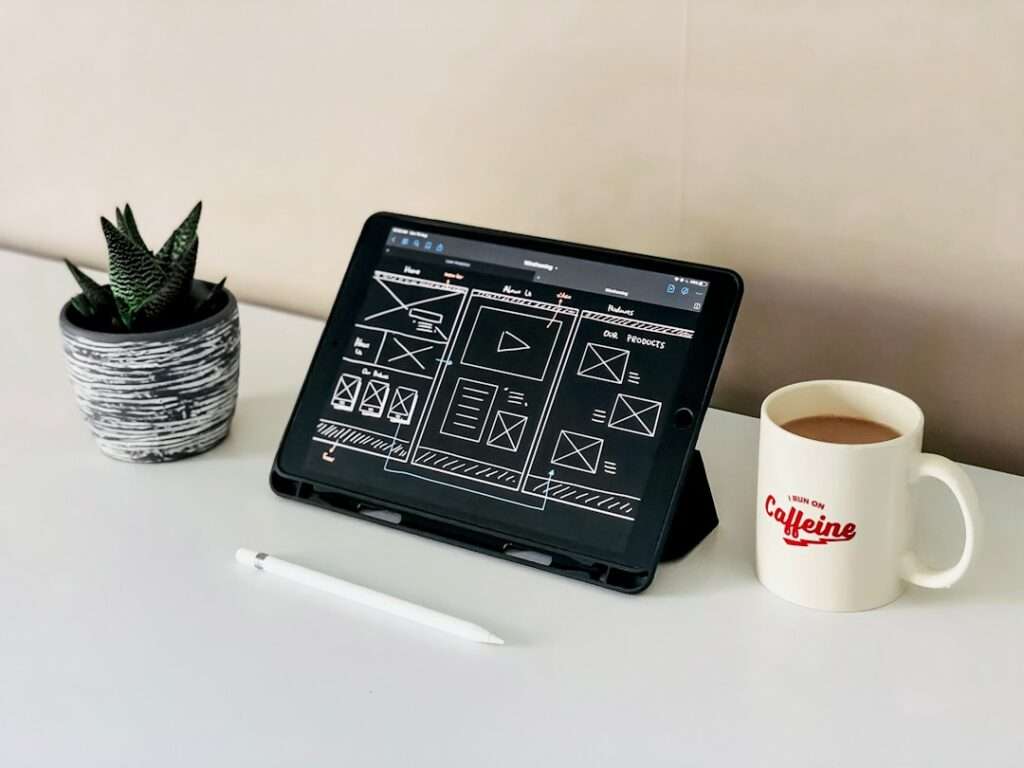 SEO Hannover - Ein Tablet mit einem Drahtgitterdesign zum Erstellen von Websites steht auf einem Ständer neben einem Stift. Eine Topfpflanze und eine Tasse Kaffee mit der Aufschrift „Koffein“ stehen ordentlich auf dem weißen Schreibtisch. Local SEO Agentur Hannover.