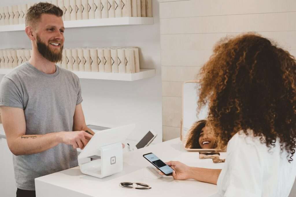 SEO Hannover - Ein Mann mit Bart und grauem T-Shirt steht hinter einem Tresen mit einem Kassenterminal und lächelt eine Frau mit lockigem Haar an, die ihr Smartphone hält. Auf dem Tresen liegt eine Sonnenbrille, die scheinbar keine Ahnung von möglichen negativen Google-Bewertungen hat. Local SEO Agentur Hannover.