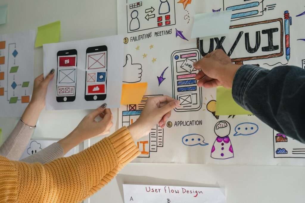 SEO Hannover - Menschen arbeiten gemeinsam an einem UI/UX-Designprojekt. Hände kleben Haftnotizen und Skizzen von mobilen Bildschirmen auf ein Whiteboard, das mit verschiedenen Zeichnungen und Diagrammen zum Benutzerfluss der App gefüllt ist. Local SEO Agentur Hannover.