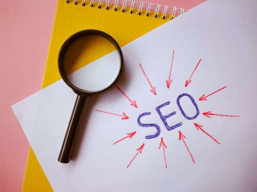 SEO Hannover - Eine Lupe liegt auf einem Stück Papier, auf dem in Lila „Google SEO“ steht, umgeben von roten Pfeilen, die auf den Text zeigen. Unter dem Papier ist teilweise ein gelbes Spiralnotizbuch sichtbar. Der Hintergrund ist rosa. Local SEO Agentur Hannover.