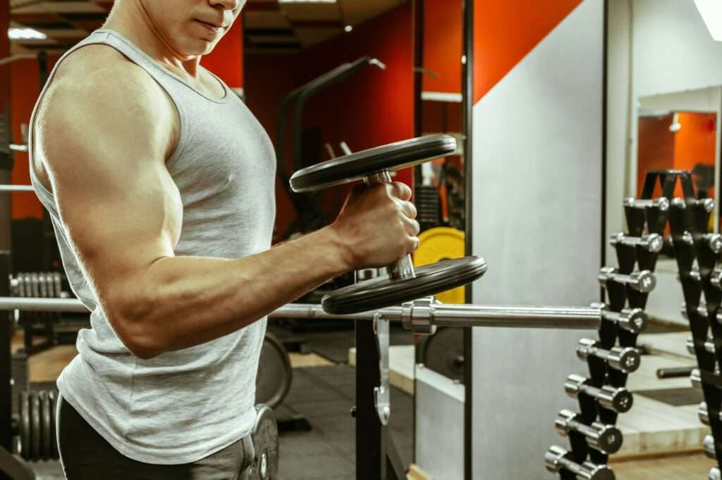 SEO Hannover - Eine Person in einem grauen Tanktop macht in einem Fitnessstudio Bizepscurls mit einer Hantel. Sie steht neben einem Hantelständer, im Hintergrund sind verschiedene Gewichte zu sehen. Das Fitnessstudio hat rot-weiße Wände. Local SEO Agentur Hannover.