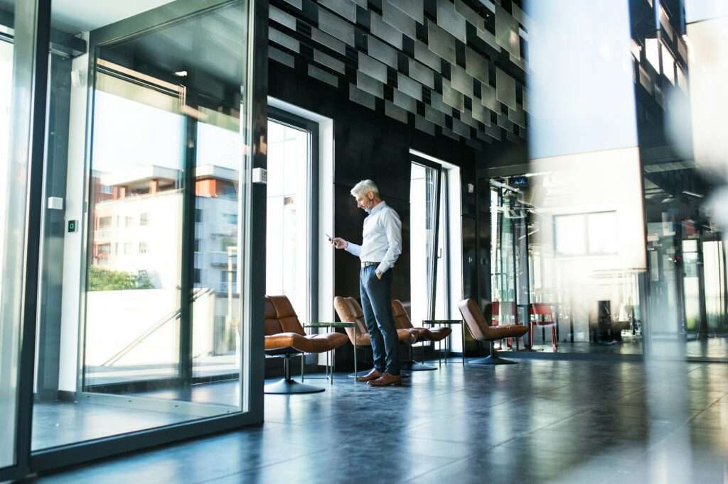 SEO Hannover - Ein Mann mit grauem Haar steht in der Lobby eines modernen Büros und schaut auf sein Telefon. Der Raum hat Glaswände, Fliesenboden und braune Lederstühle. Local SEO Agentur Hannover.