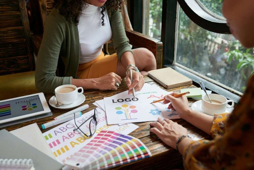 SEO Hannover - Zwei Personen sitzen an einem Holztisch mit Notizbüchern, Kaffeetassen, Farbmustern und Materialien von Designer für Logos. Eine zeigt auf ein gedrucktes Logo, während die andere einen Stift hält. Durch das große Fenster neben ihnen fällt Tageslicht ein. Local SEO Agentur Hannover.