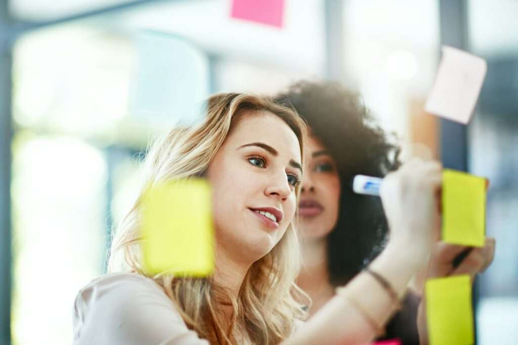 SEO Hannover - Zwei Personen arbeiten in einem Büro zusammen und benutzen bunte Haftnotizen an einer Glaswand. Einer schreibt mit einem Marker, während der andere, von einer SEO Agentur Hannover, zusieht. Die Szene suggeriert Teamarbeit und Brainstorming in einem modernen Arbeitsumfeld. Local SEO Agentur Hannover.