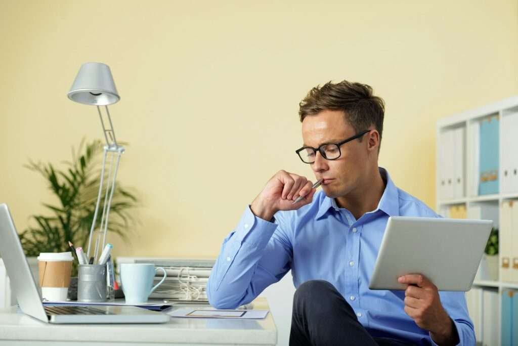 SEO Hannover - Ein Webdesigner aus Hannover sitzt mit Brille an einem Schreibtisch in einem Büro, hält ein Tablet in der Hand und legt nachdenklich die Hand auf sein Kinn. Auf dem Schreibtisch stehen ein Laptop, eine Lampe, eine Kaffeetasse und Ordner. Eine Pflanze und Regale mit weiteren Ordnern vervollständigen den kreativen Arbeitsplatz. Local SEO Agentur Hannover.