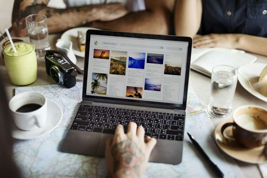 SEO Hannover - Eine Person mit Tattoos auf der Hand benutzt einen Laptop, auf dem Bilder verschiedener Landschaften angezeigt werden, wahrscheinlich für ein Webvisitenkarte-Projekt. Auf dem Tisch stehen Kaffee, ein Smoothie, eine Kamera und Wassergläser. Im Hintergrund sind zwei weitere Personen teilweise zu sehen. Local SEO Agentur Hannover.