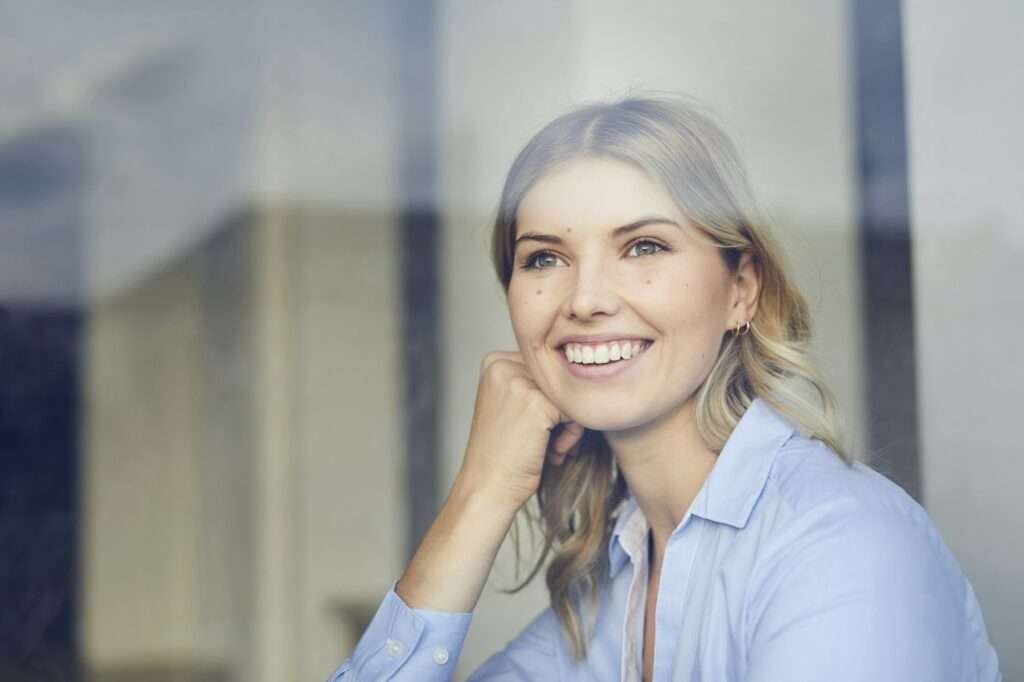 SEO Hannover - Eine Frau mit blonden Haaren und hellblauem Hemd lächelt und blickt nachdenklich an ein Bürofenster. Sie verkörpert den Innovationsgeist einer SEO-Agentur in Hannover. Sie stützt ihr Kinn auf ihre Hand und ist in Gedanken versunken, während Sonnenlicht die kreative Atmosphäre des Raums erhellt. Local SEO Agentur Hannover.