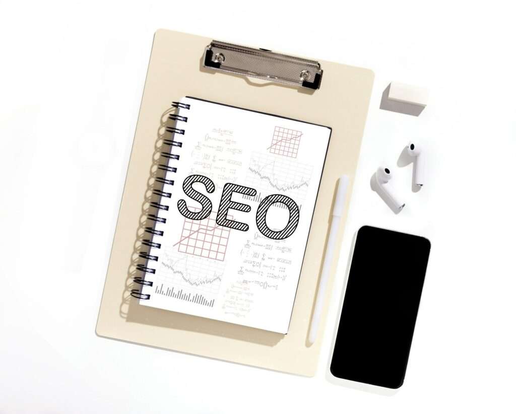 SEO Hannover - Auf einem Klemmbrett liegt ein Spiralnotizbuch mit der Aufschrift „SEO“ auf dem Einband, umgeben von Diagrammen und Kritzeleien. Daneben liegen ein Smartphone, kabellose Ohrhörer, ein Stift und ein Radiergummi, alles auf einer weißen Oberfläche angeordnet – die perfekte Umgebung für eine Brainstorming-Sitzung der SEO Agentur Hannover. Local SEO Agentur Hannover.