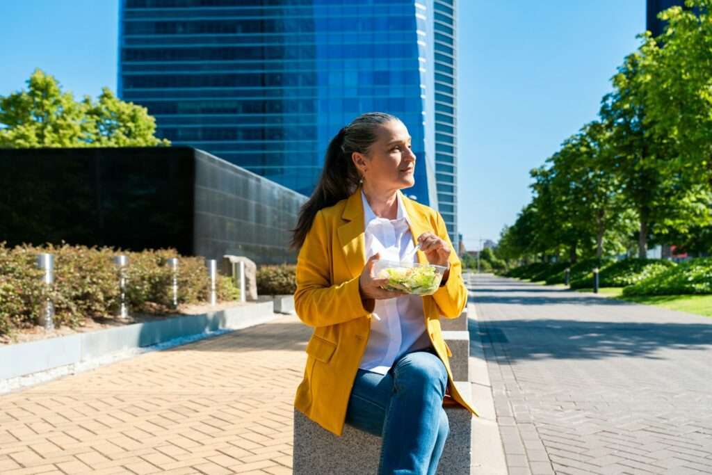 SEO Hannover - Eine Person in einem gelben Blazer sitzt auf einer Betonbank und genießt eine Schüssel Salat. Dieses malerische Foto fängt die Harmonie von Natur und Stadtleben ein, mit einem von Bäumen gesäumten Weg, der zu einem hohen Gebäude unter der warmen Umarmung der Sonne führt. Local SEO Agentur Hannover.