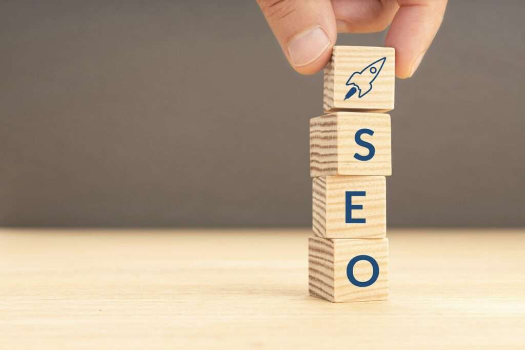 SEO Hannover - Ein Stapel Holzklötze bildet das Wort „SEO“ mit einem Raketensymbol auf dem obersten Klotz, das den Start Ihrer Google SEO-Strategie symbolisiert. Eine Hand legt den obersten Klotz vorsichtig auf den Stapel vor einem neutralen Hintergrund, bereit, Ihre Online-Präsenz auf neue Höhen zu katapultieren. Local SEO Agentur Hannover.