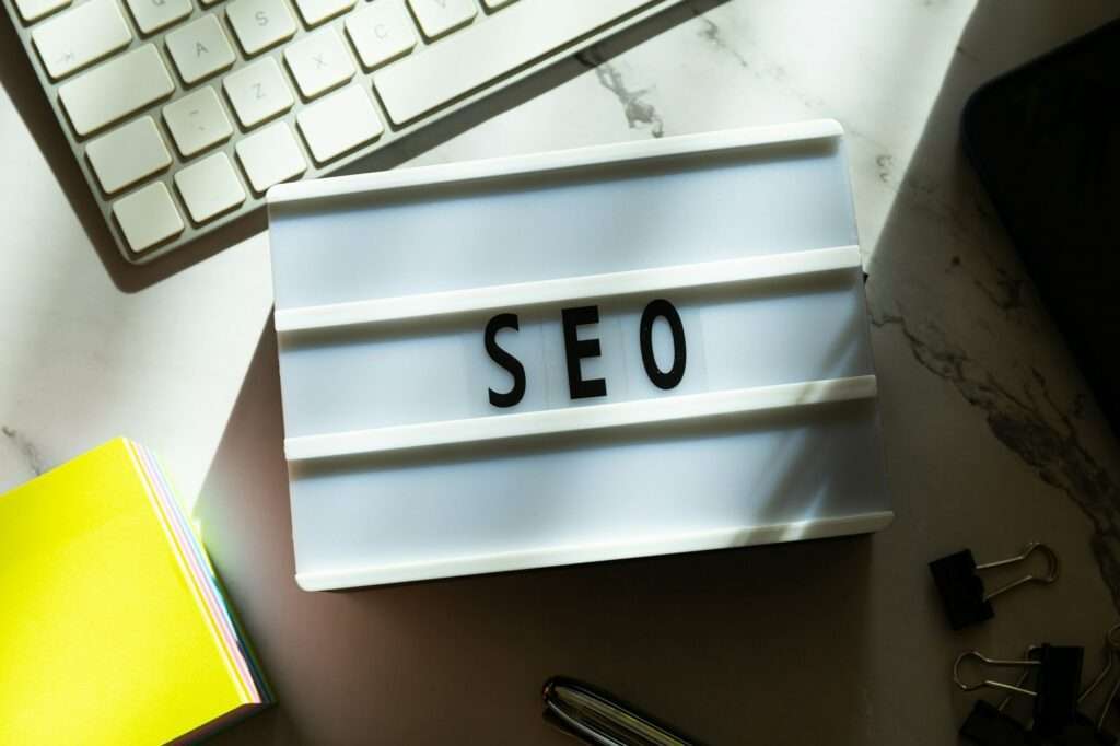 SEO Hannover - Auf einem Schreibtisch steht ein Leuchtkasten mit dem Wort „Google SEO“. Daneben stehen eine Computertastatur, ein gelbes Notizbuch, Heftklammern und ein Stift, alles im Tageslicht. Local SEO Agentur Hannover.