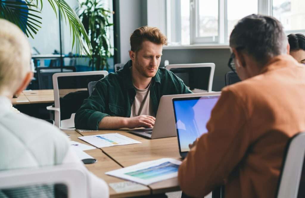 SEO Hannover - Eine Gruppe von Menschen sitzt an einem Tisch und arbeitet an Laptops. Auf dem Tisch liegen verstreut Papiere mit Diagrammen, Grafiken und Notizen zu „Kosten für Website-Erstellung“. Sonnenlicht fällt durch große Fenster herein und wirft einen warmen Schein auf das Grün im Hintergrund. Local SEO Agentur Hannover.