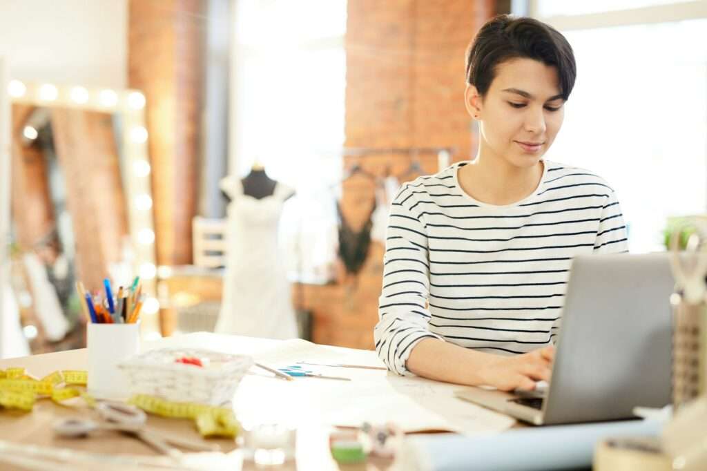 SEO Hannover - Eine Person im gestreiften Hemd arbeitet vertieft an ihrem Laptop an ihrem Schreibtisch in einem Modestudio. Zwischen Nähutensilien und Skizzen schimmert das Wesen eines Kunden der Webagentur Hannover hervor. Im Hintergrund ziert ein Kleid eine Schaufensterpuppe im sanften Licht der Schminktischlampen. Local SEO Agentur Hannover.